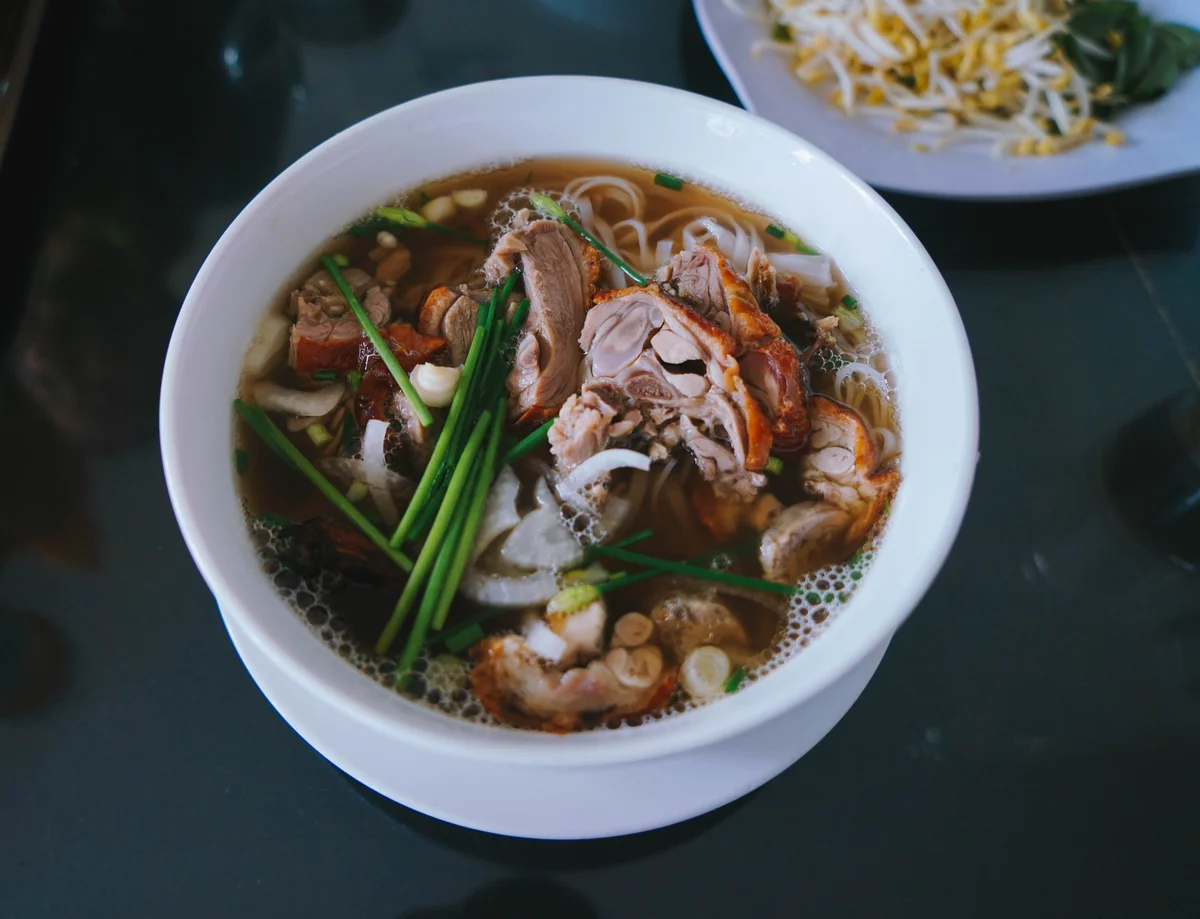 Bowl of noodle soup with sliced meat, green onions, and broth on a dark table. Plate of bean sprouts and greens in the background.