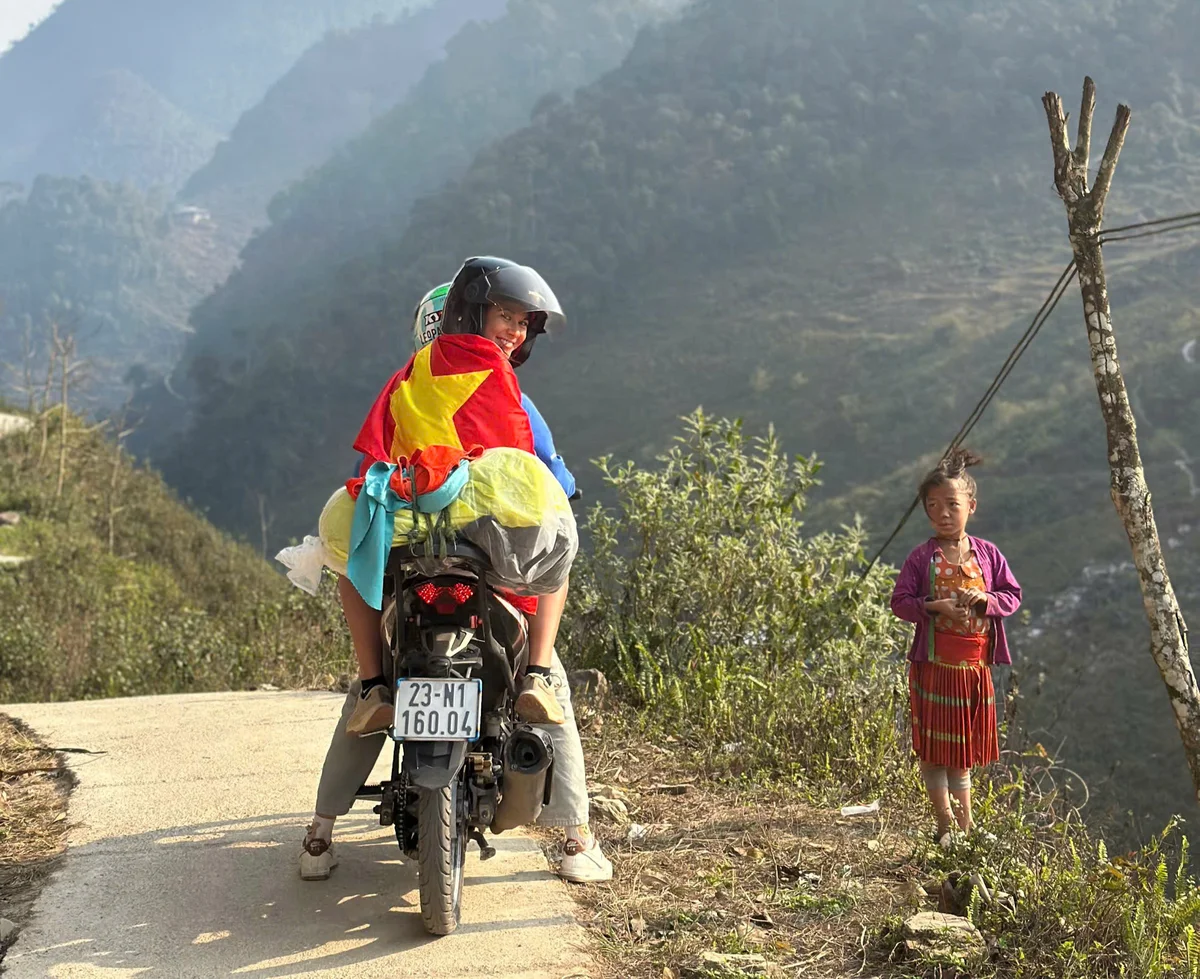 Two people on a motorcycle with a red-yellow flag, smile on a mountain road. A child in traditional attire stands nearby. Sunny, green hills.