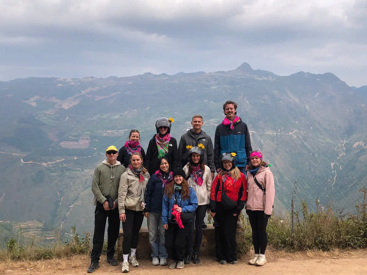 Group of people posing with colorful scarves and flowers, overlooking a mountainous landscape under a cloudy sky. Peaceful mood.