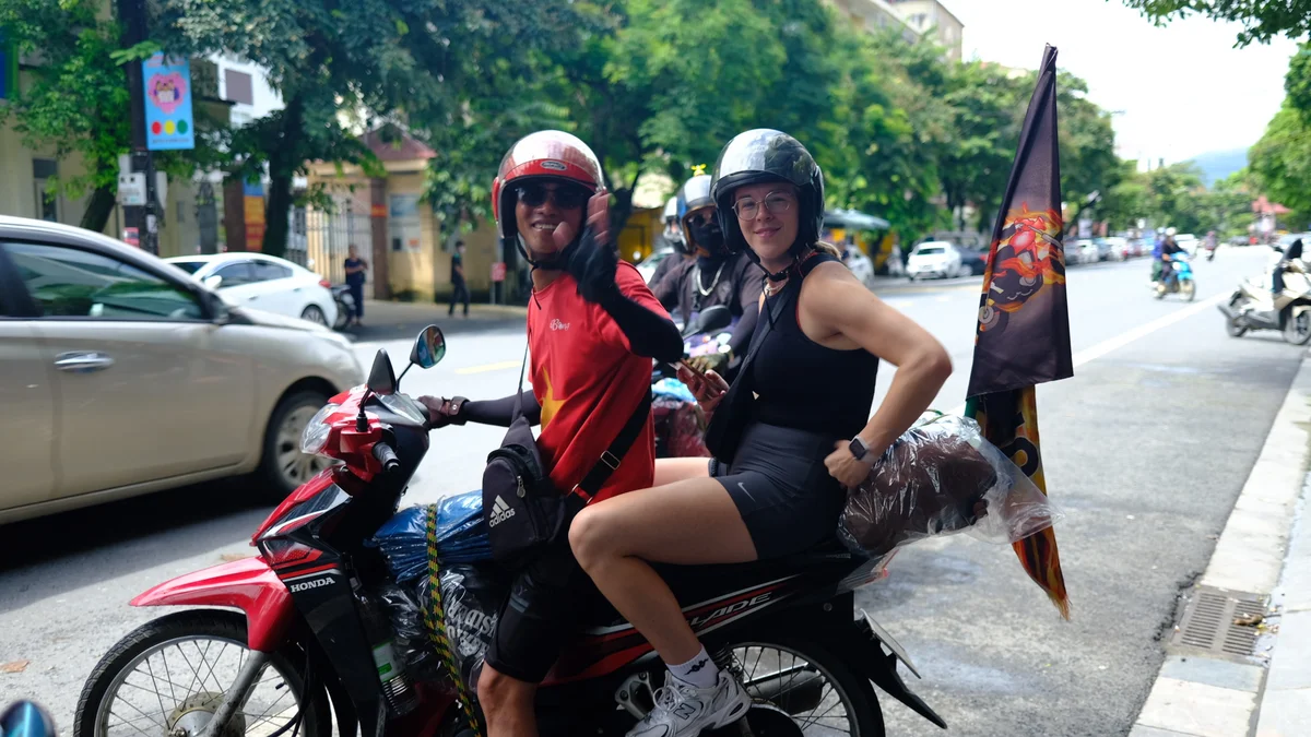 Two people on a red motorbike, waving and smiling, wearing helmets. Street with cars and trees in the background. Casual, cheerful vibe.