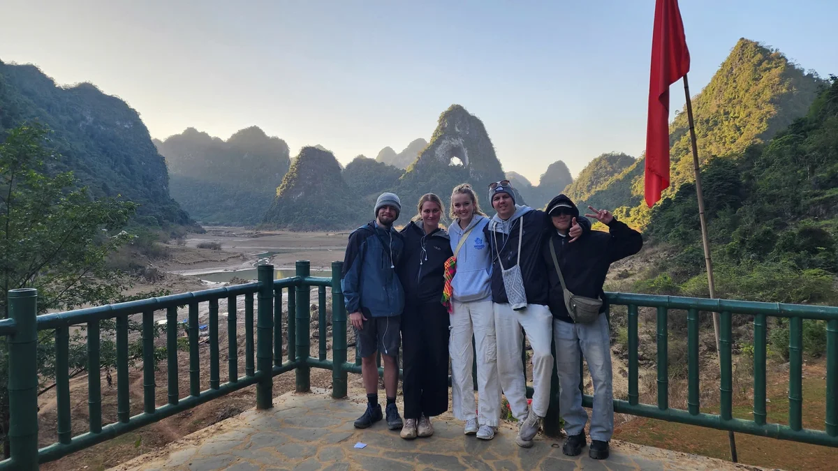 Five people smiling, posing on a railing with scenic mountain backdrop. Red flag on right. Clear sky, relaxed mood.