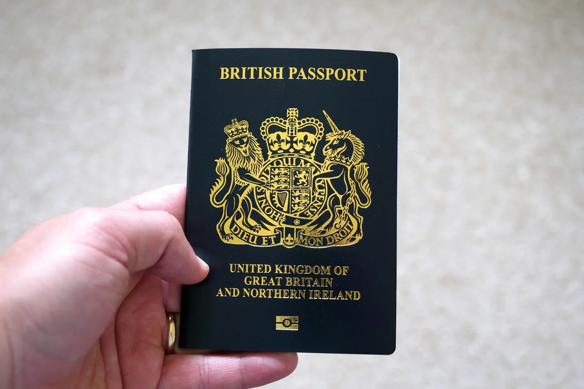 A hand holds a black British passport with a gold crest and text "United Kingdom of Great Britain and Northern Ireland" against beige background.