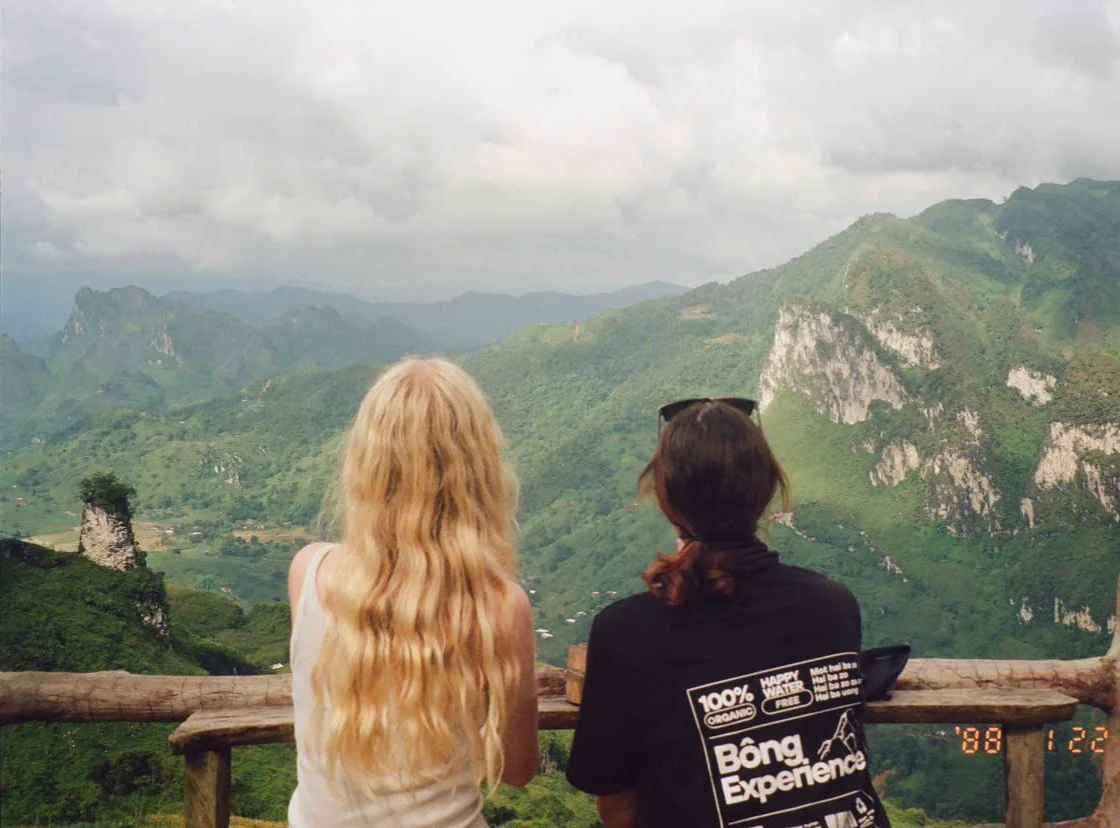 Two people with long hair admire a vast mountainous landscape from a wooden railing. One wears a shirt with printed text. The mood is serene.