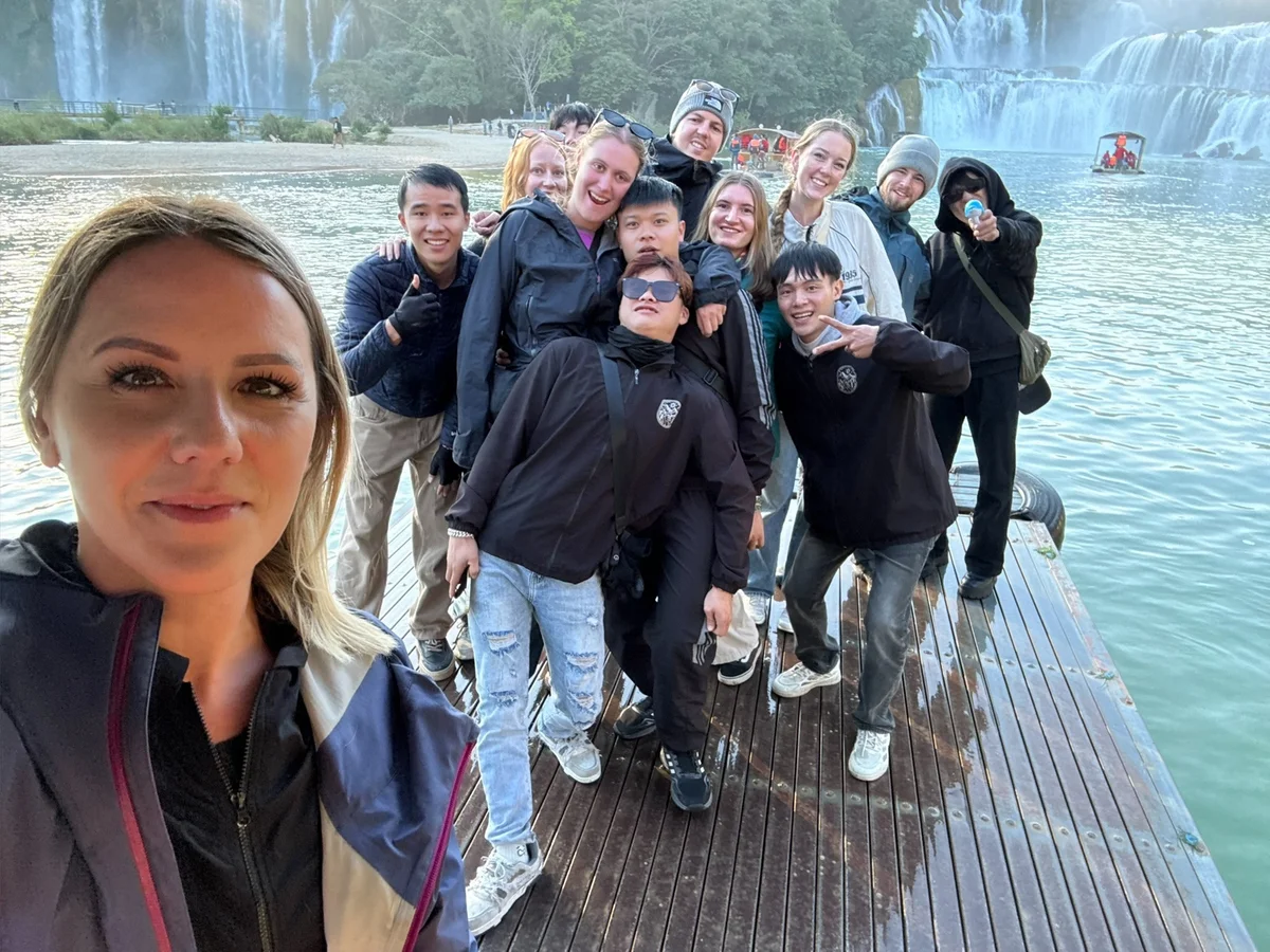 Group posing on a dock in front of waterfalls, with water and trees in the background. Everyone is smiling, creating a joyful atmosphere.