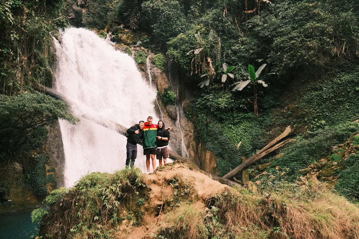 Three people smiling in front of a waterfall surrounded by lush greenery. They stand on a mossy hill, exuding a cheerful, adventurous vibe.