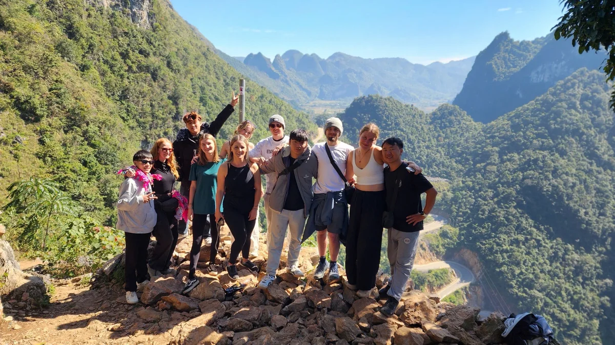 Group of people smiling and posing on a rocky cliff with lush green mountains in the background under a clear blue sky.