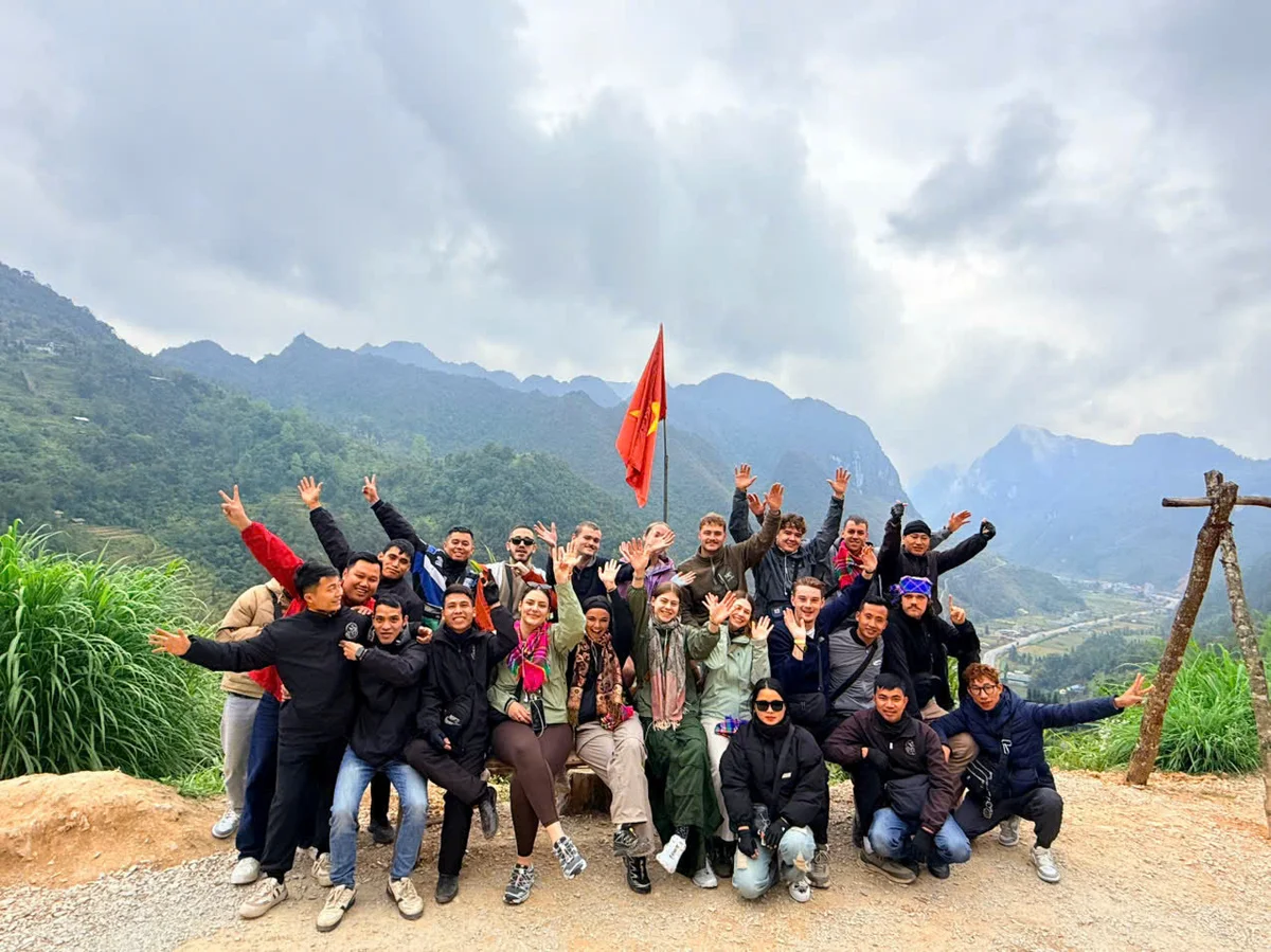 Group of people posing cheerfully with raised arms, red flag in background, set in a mountainous landscape under a cloudy sky.