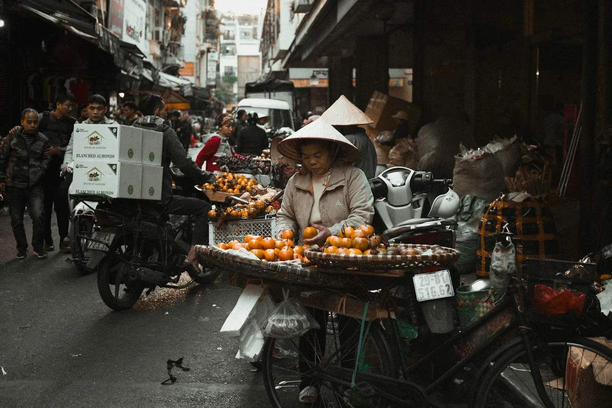 Street market scene with a vendor in a conical hat arranging oranges on a bicycle. Crowded background with people and motorbikes.