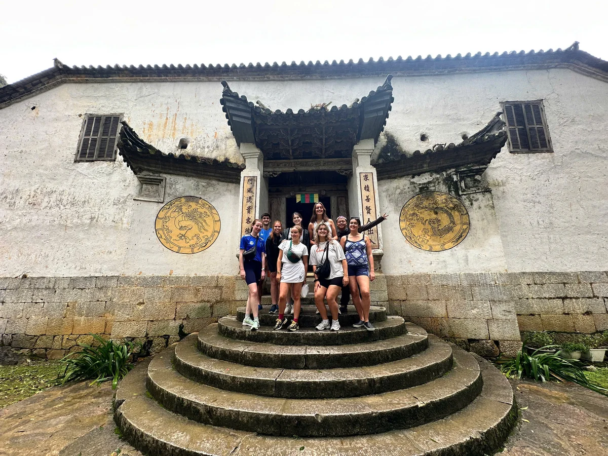A group of people smiling on stone steps in front of an ancient building with ornate carvings and large circular engravings.