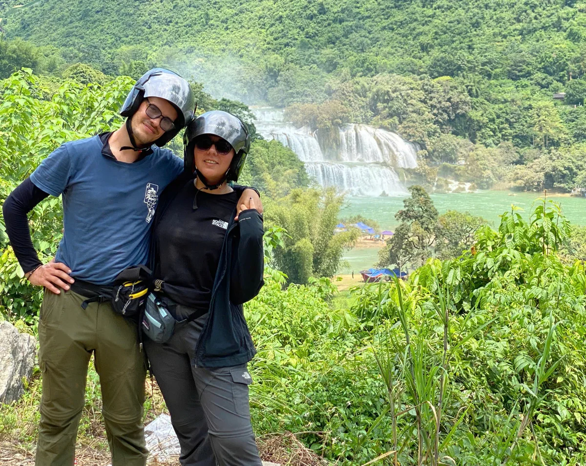 Two people in helmets pose in front of a lush green landscape with a waterfall. They are smiling, exuding a cheerful mood.