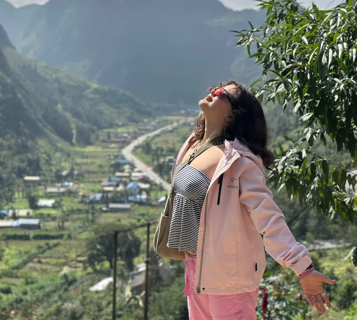 Woman in a pink jacket and striped top joyfully stretches arms out in a mountainous valley. Sunlight and greenery create a vibrant scene.