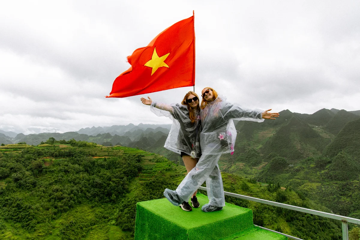 Two people in rain ponchos pose joyfully atop a green platform with a red flag against a backdrop of lush green mountains and cloudy sky.