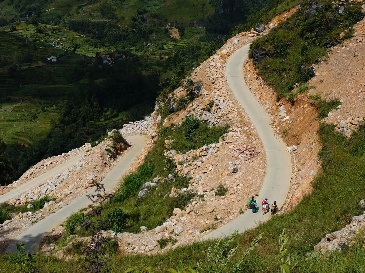 Three cyclists ride uphill on a winding mountain road surrounded by greenery and rocks, under a clear, sunny sky.