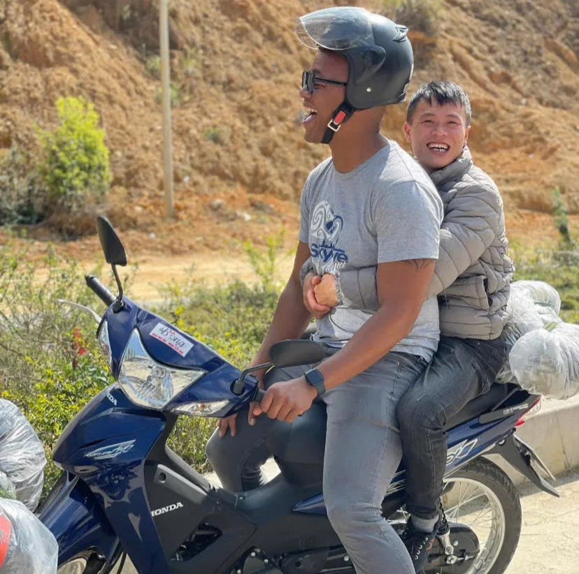 Two people laughing on a blue motorbike, helmeted driver in a t-shirt, passenger hugging from behind. Sunlit, earthy background.