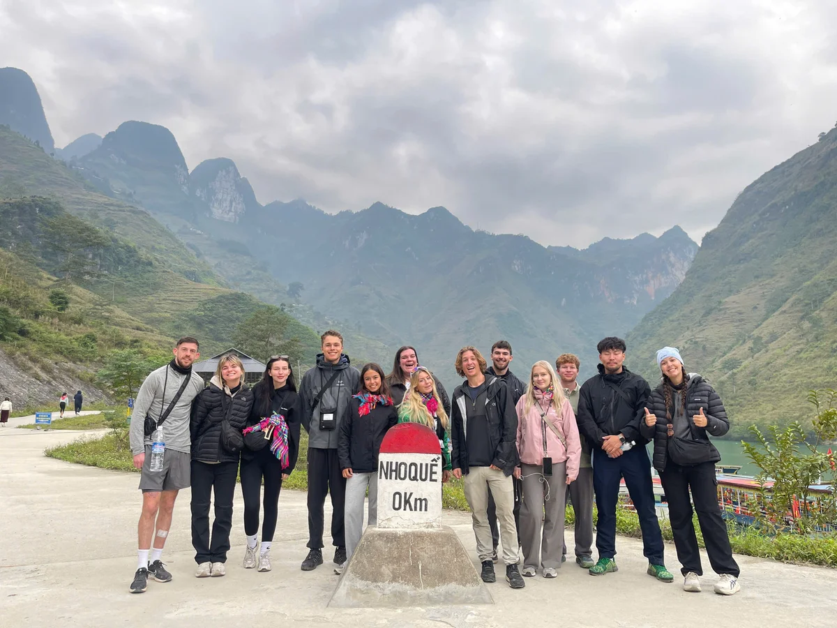 Group of people smiling by a "Nho Que 0Km" marker in a mountainous landscape, overcast sky. Some wear jackets and winter hats.