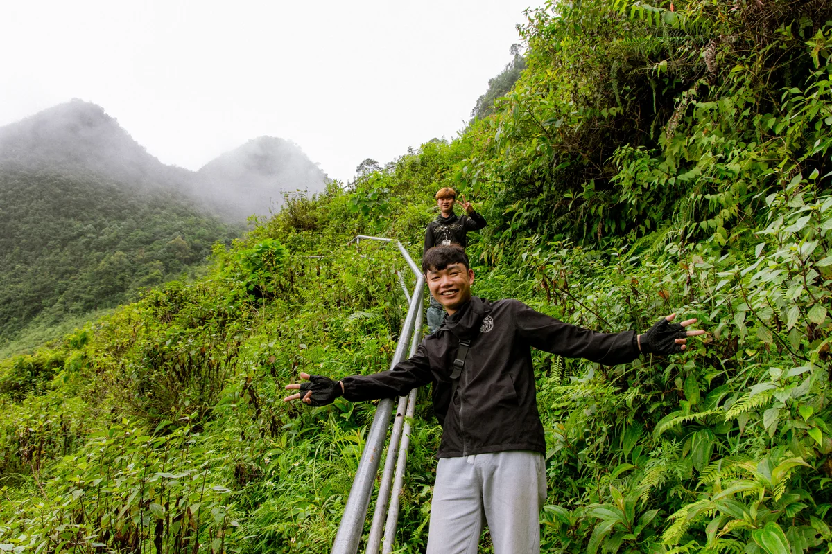 Two people on a lush green mountain trail, smiling and posing with peace signs. Misty hills in the background create a lively mood.