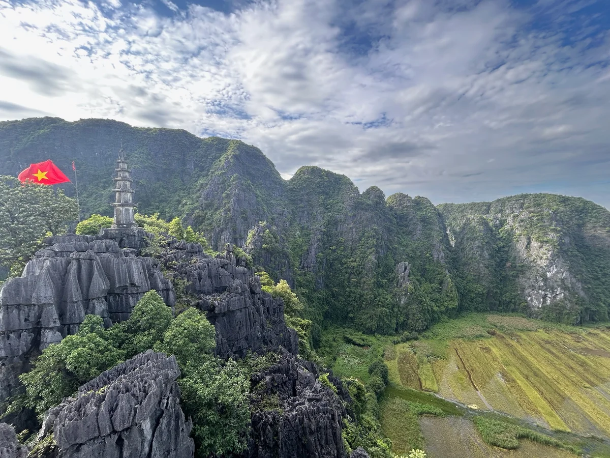 Rocky hill with a pagoda and a Vietnamese flag under a cloudy sky, surrounded by lush green mountains and a rice field in the valley.