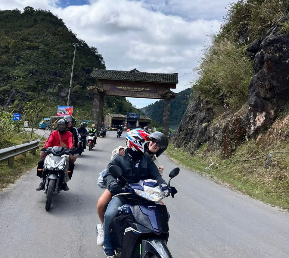 Motorcyclists ride through a scenic mountain road under a stone arch. Two people in front share a joyful moment. Riders wear helmets.