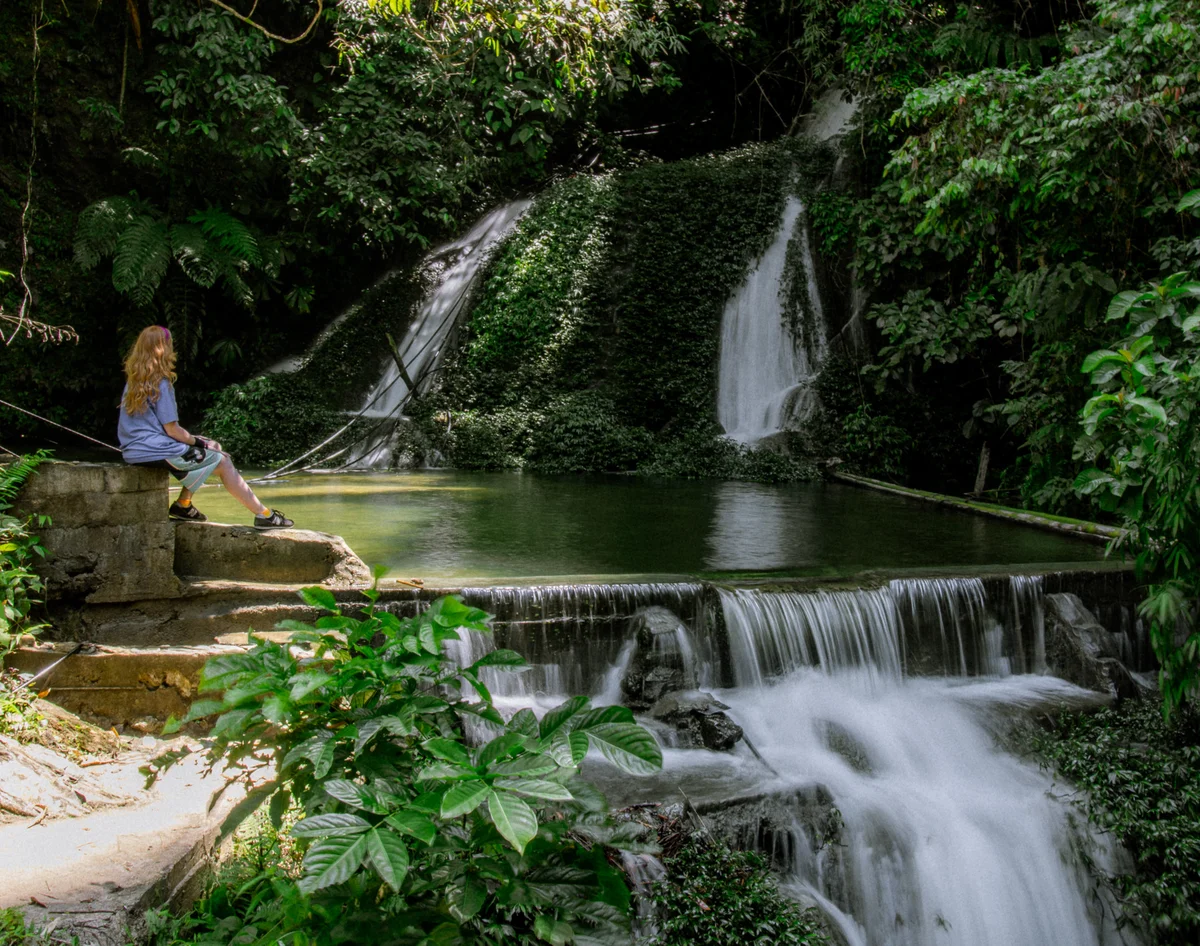 A person with long hair sits by steps near a lush waterfall and pool, surrounded by greenery. The mood is tranquil and serene.