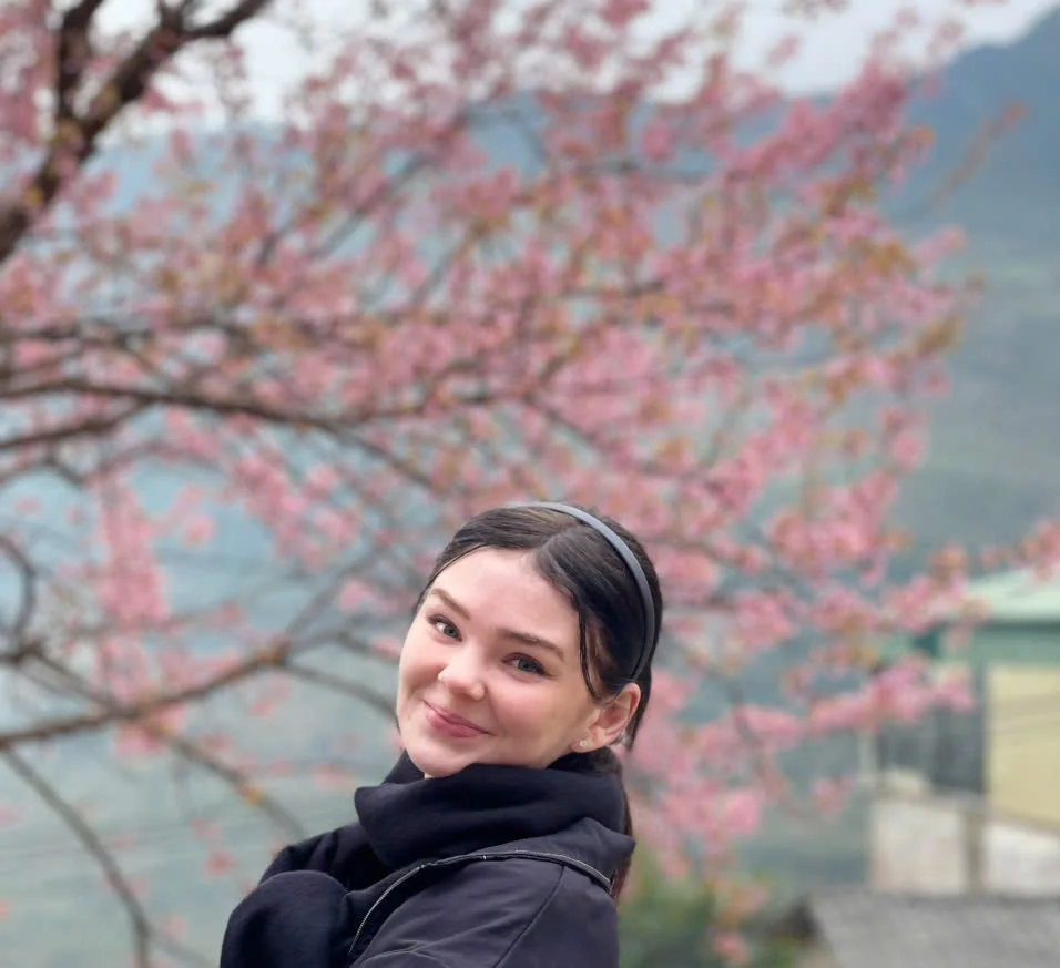 A smiling woman in a black jacket and headband stands in front of blooming pink cherry blossoms with a misty mountain background.
