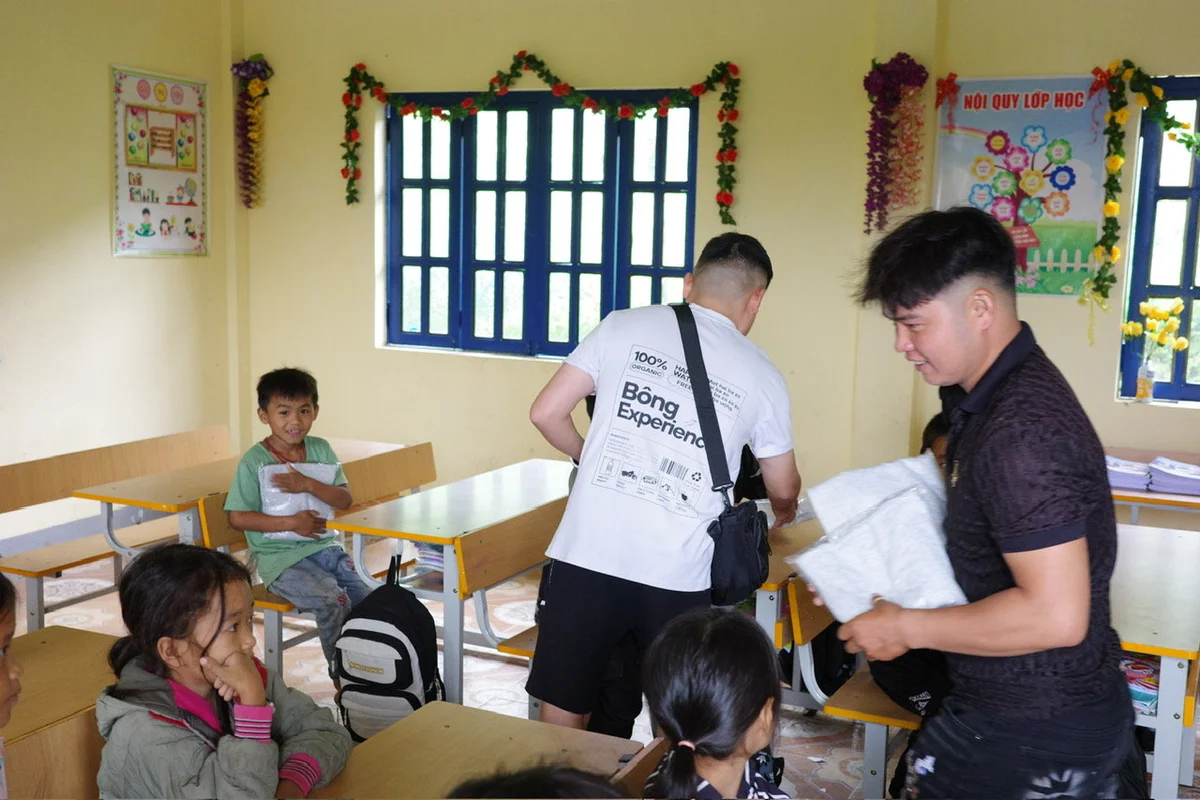 Children in a classroom receive gifts from two men. A smiling boy holds a package. Decorated walls and windows in the background indicate a cheerful setting.
