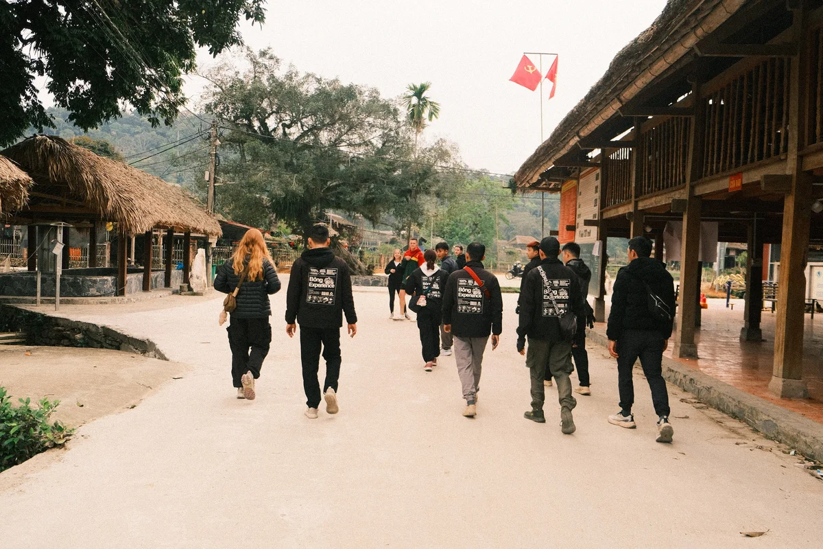 A group of people in dark jackets walk through a rustic village with thatched huts and a Communist flag. The mood is casual and serene.