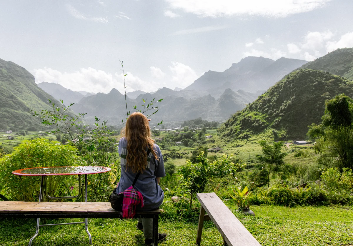 A woman with long hair sits on a bench facing lush green hills under a cloudy sky. A table and a bag are nearby, creating a serene mood.