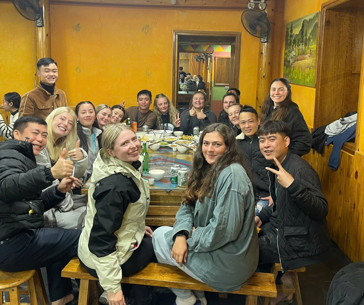 A group of people smiling and posing around a wooden table with food and drinks in a cozy room with orange walls, exuding a joyful mood.