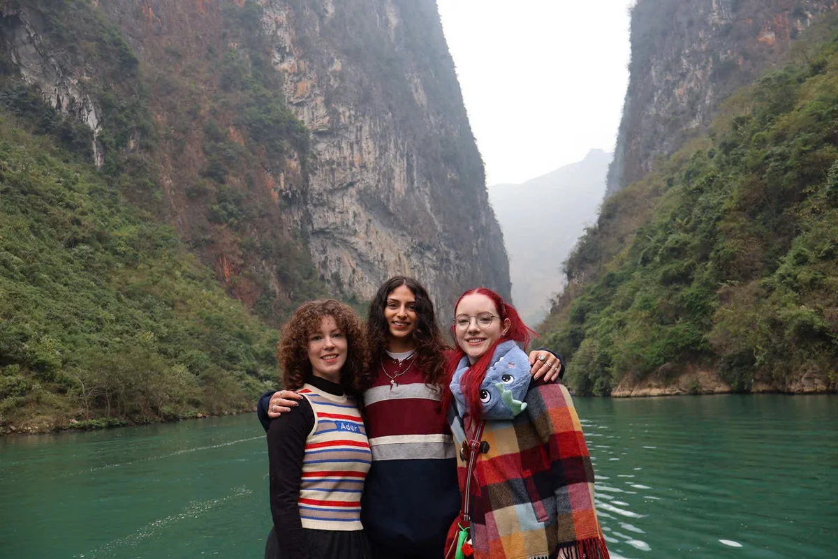 Three people smile near a turquoise river, surrounded by tall green cliffs. One wears a colorful striped sweater, another has a bold red checkered scarf.