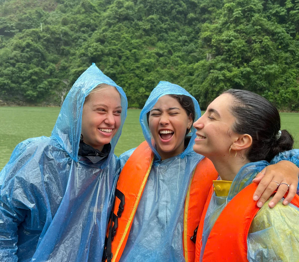 Three people in blue rain ponchos and orange life vests laugh together. They're outside near lush greenery and water, creating a joyful mood.