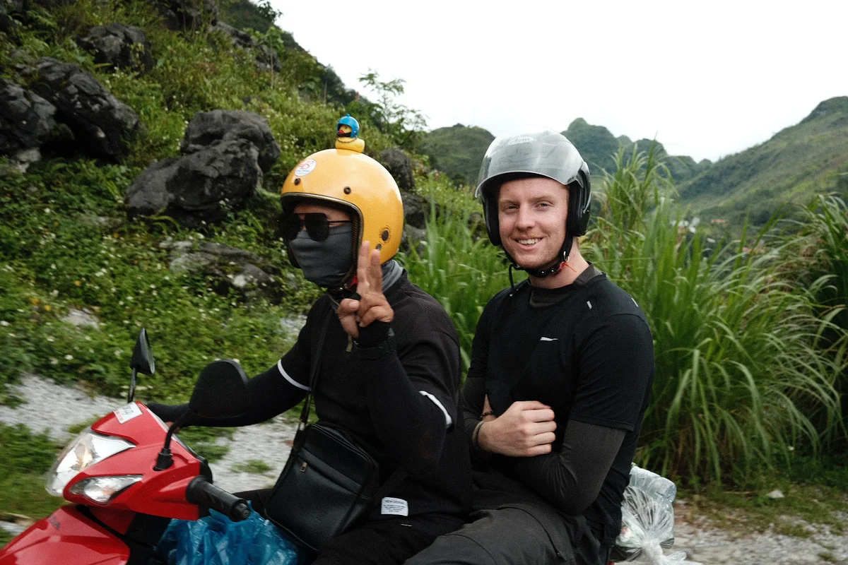 Two people riding a red scooter in a green mountainous area. One wears a helmet with a toy duck, making a peace sign, the other smiles.