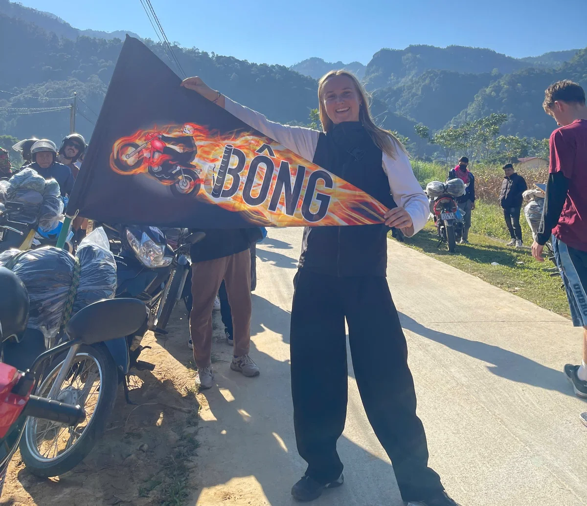Smiling person holds a triangular flag with "BÔNG" and fiery motorcycle design. Background of motorcycles and hills under a clear sky.