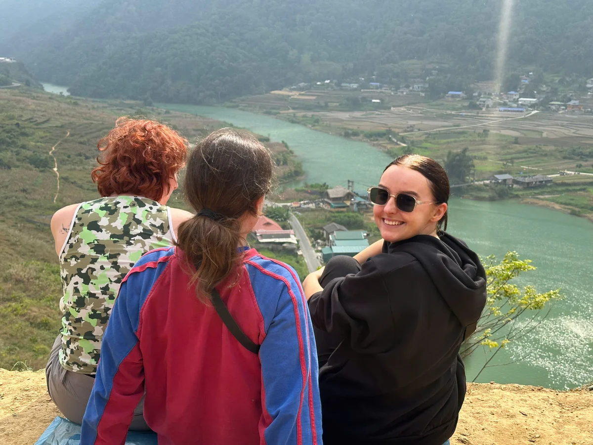 Three people sit on a hill overlooking a river and village. The woman in sunglasses smiles, wearing a black jacket. The mood is relaxed.