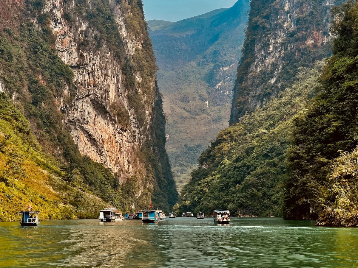 Boats float on a green river between towering cliffs in a lush canyon. The scene is serene, with mountains under a clear blue sky.