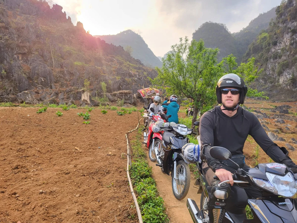 People on motorcycles ride through a rocky path in a mountainous landscape. The setting sun casts warm light, highlighting greenery and hills.