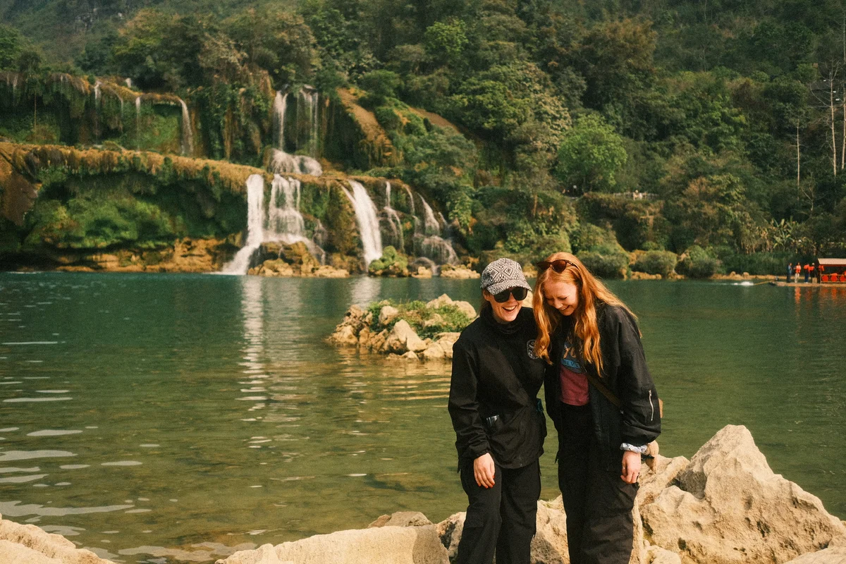 Two people in black jackets smile near a lake with a waterfall and lush green landscape. Clear water and rocks in the foreground.