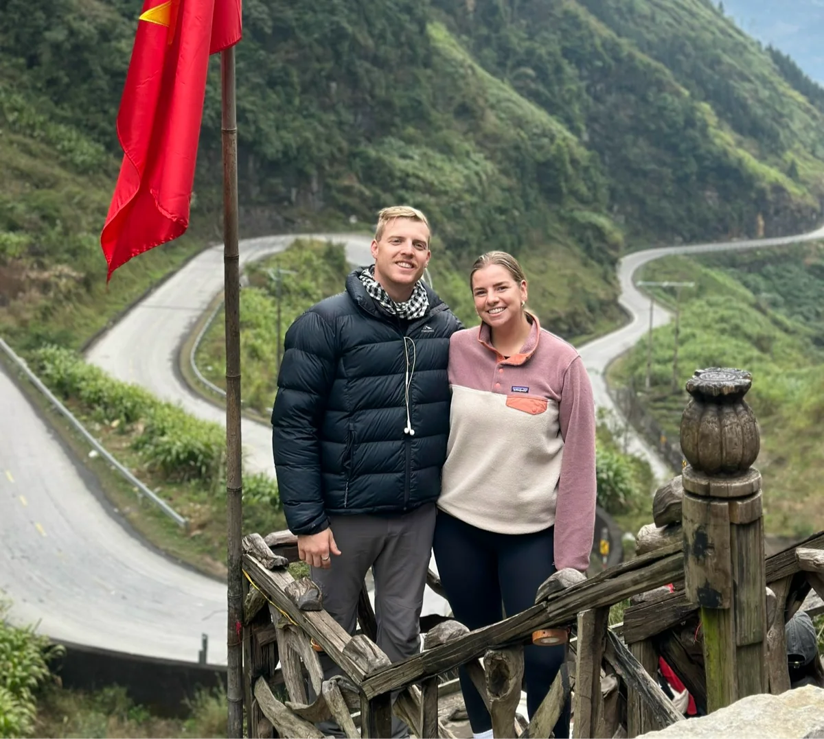 Two people smiling, standing on a wooden platform with a winding road and green hills in the background. A red flag is visible.