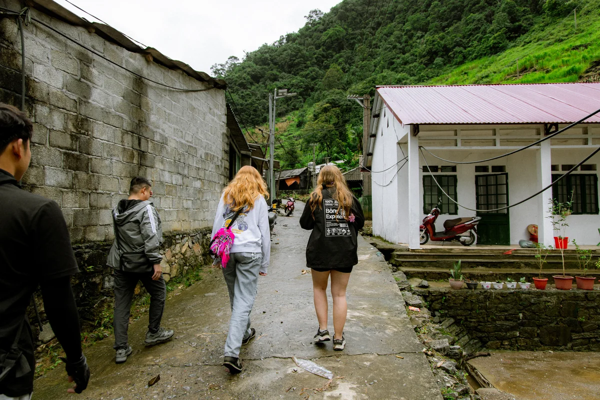 People walking on a village path flanked by stone and white buildings, red roof and lush green hill in background, carrying bags.