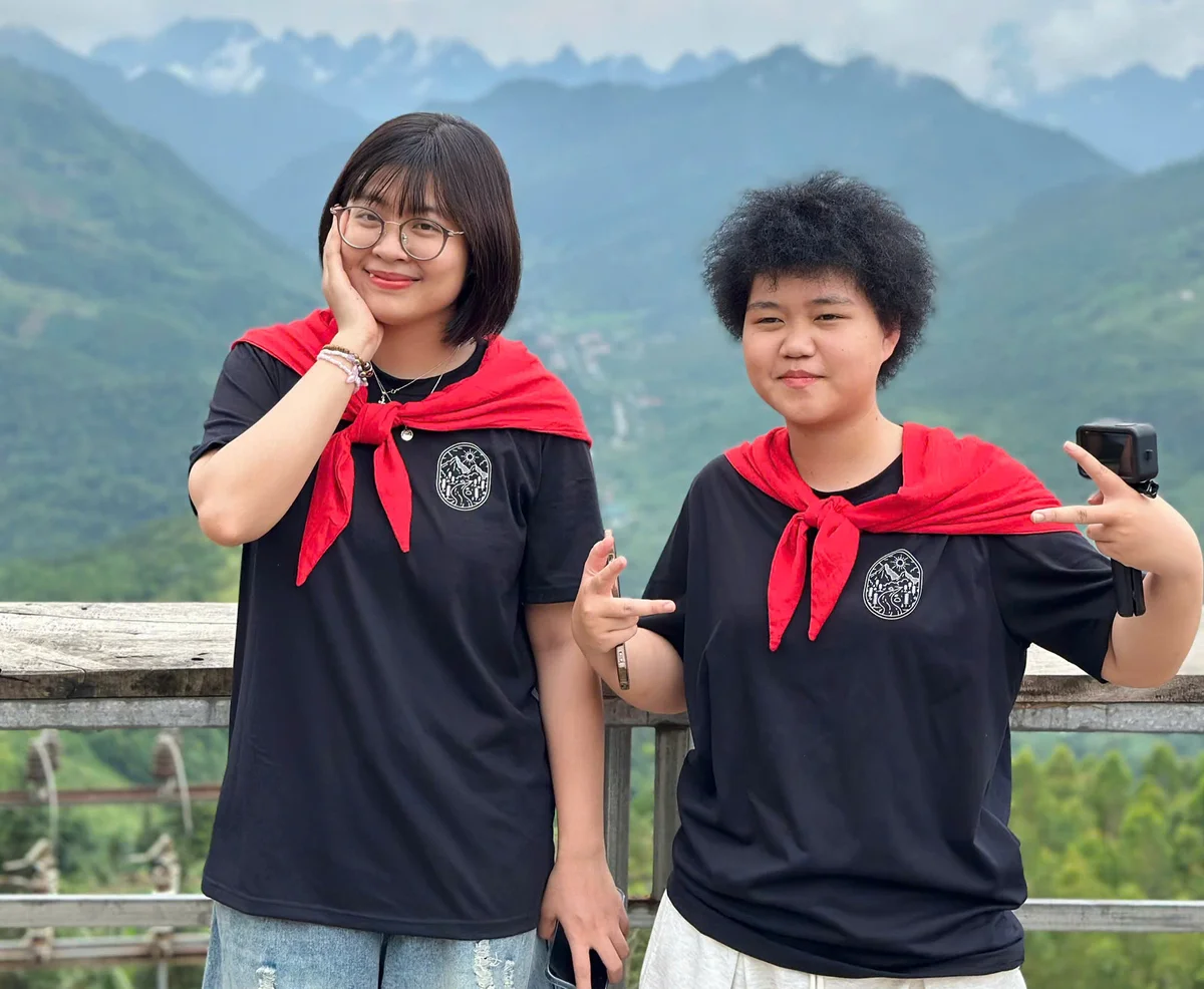 Two people in black shirts with red scarves pose on a mountain balcony. One smiles, the other makes peace signs. Cloudy sky and green hills.