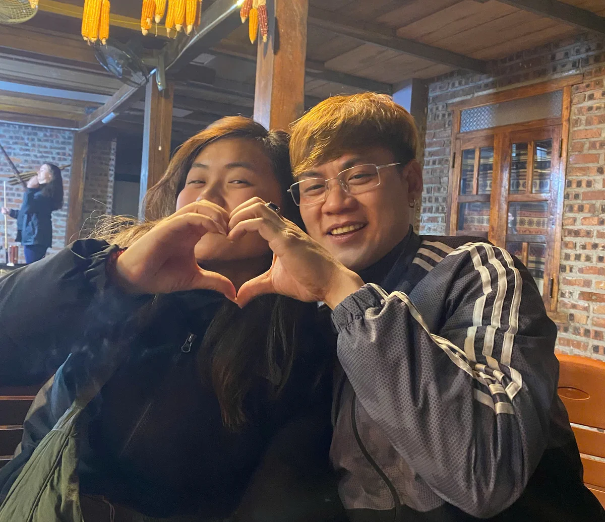 Two people form a heart with their hands, smiling in a cozy, wooden interior. Corn hangs above; a brick wall and warm light set a friendly mood.