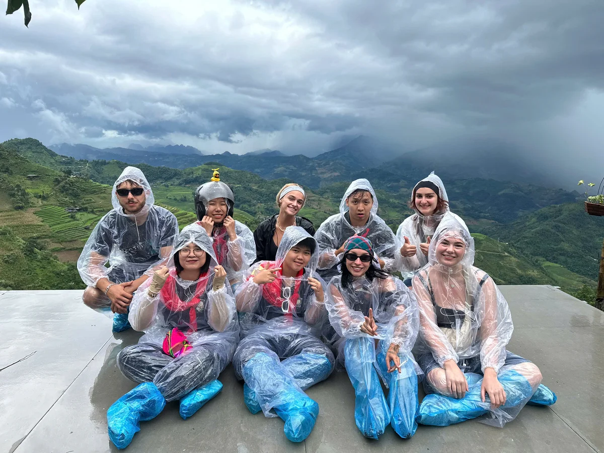 A group of people in clear rain ponchos sit and smile with thumbs up on a mountain overlook. The sky is cloudy, and the mood is cheerful.