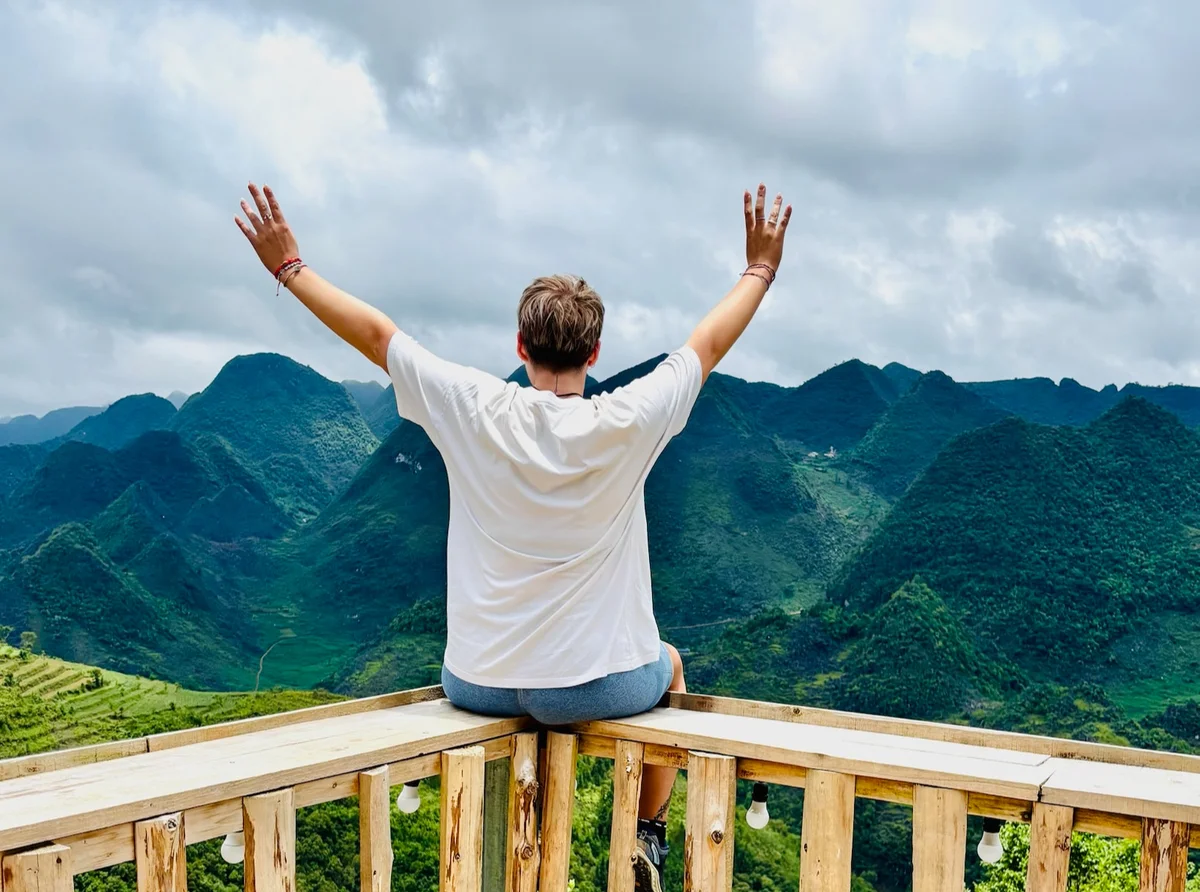 Person in a white shirt sits on a wooden railing with arms raised, overlooking lush green mountains under a cloudy sky, exuding freedom.