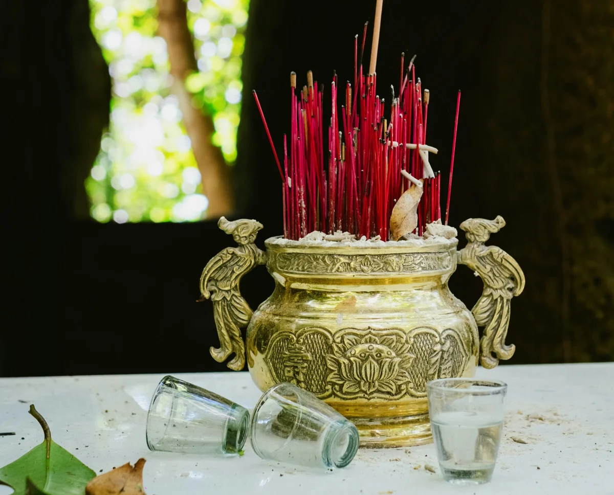 Golden incense holder with red sticks on a white table, two empty glasses, and blurred green foliage in the background. Calm atmosphere.