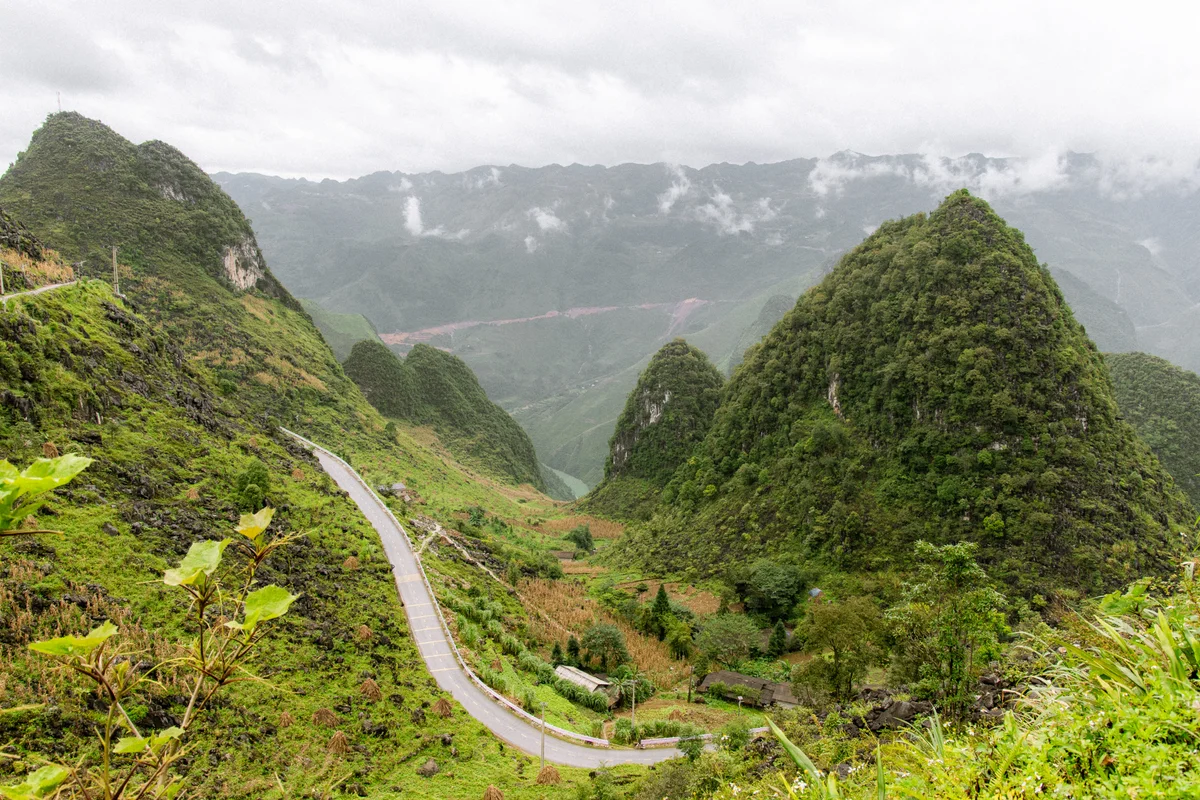 Winding road through lush green mountains under a cloudy sky. Verdant landscape with steep peaks, trees, and low mist creates a serene mood.