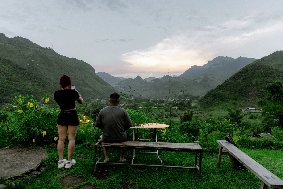 Person takes photo of mountains at sunset; another sits on bench in lush garden. Yellow flowers, green hills, and cloudy sky visible.