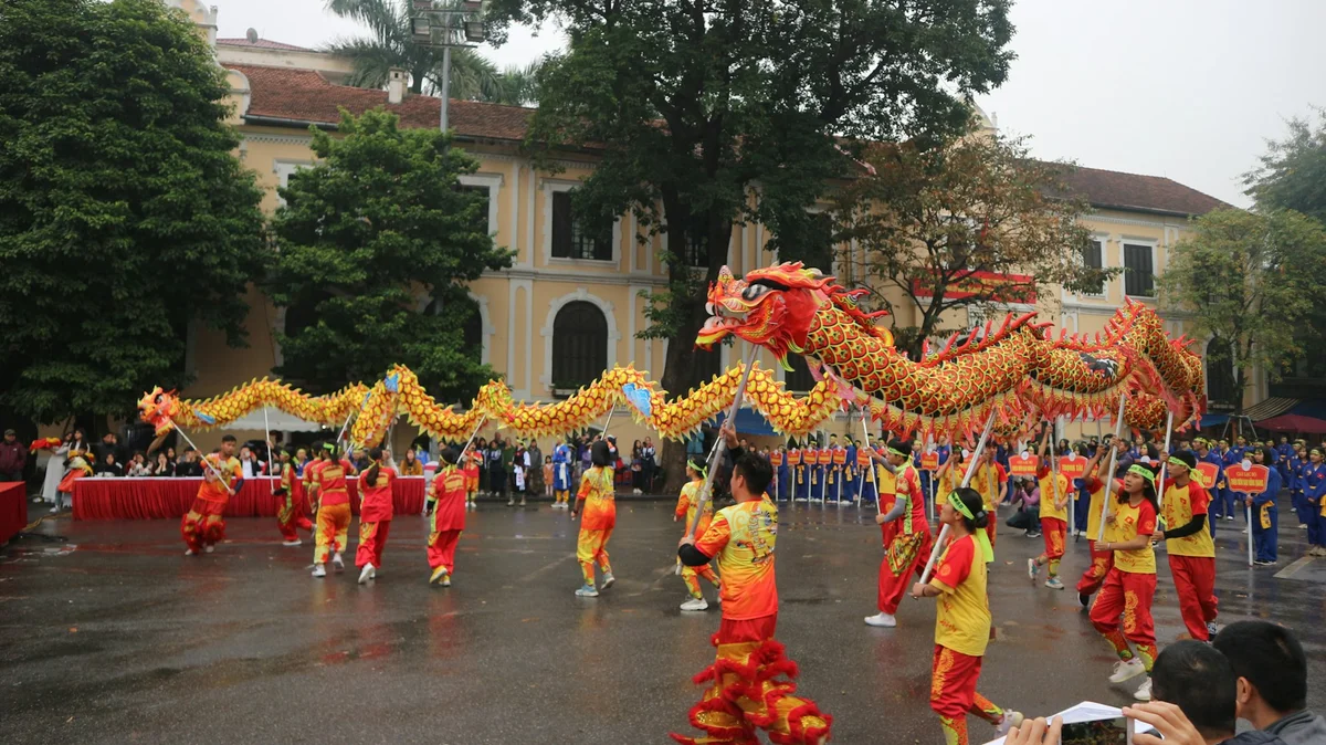Performers in vibrant red and yellow costumes dance with a dragon puppet in a lively outdoor festival, with a crowd and historic buildings in the background.