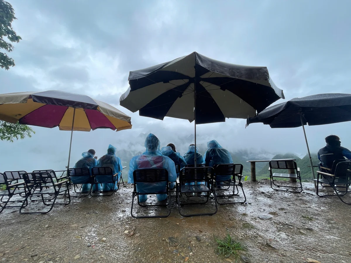People in blue raincoats sit under umbrellas, overlooking a foggy mountain view. The scene is wet and overcast, creating a serene mood.
