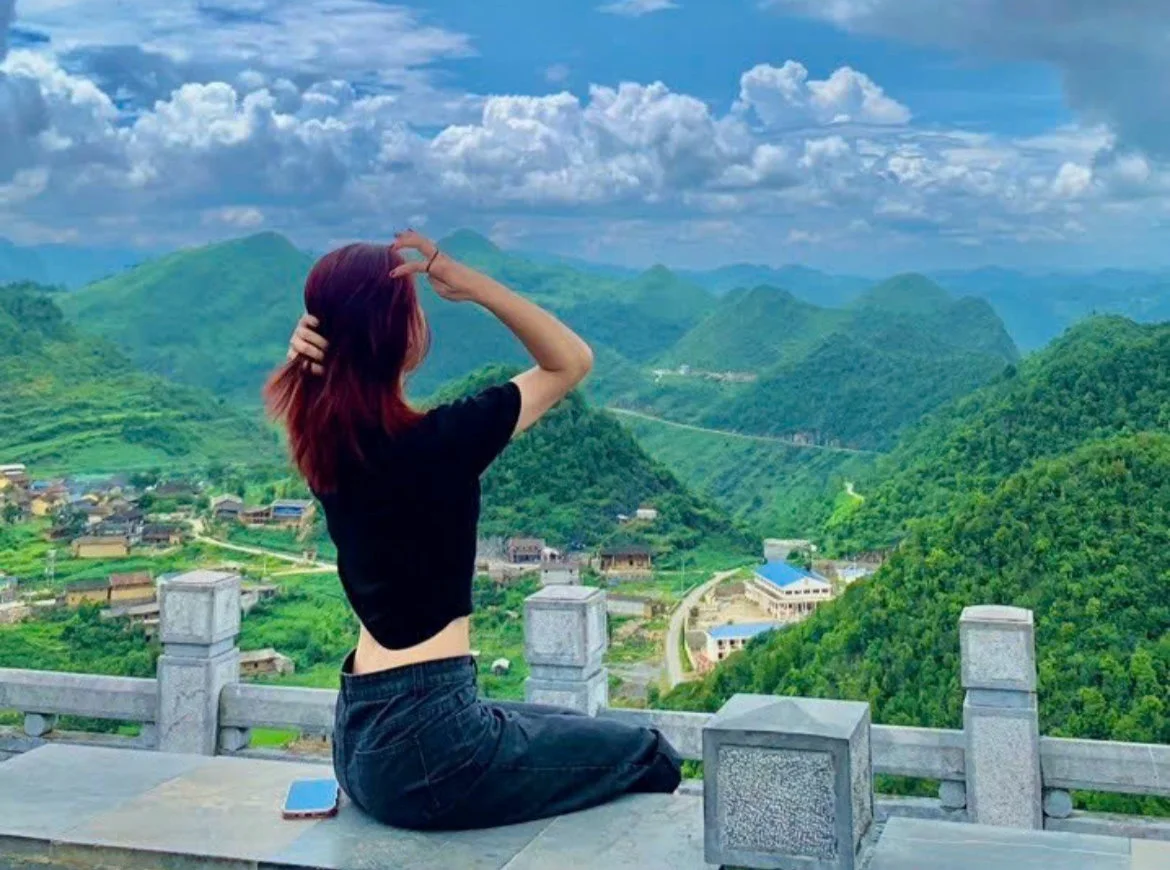 Woman in black top sits on a stone railing, looking at lush green mountains and a cloudy sky. A phone lies beside her on the ledge.