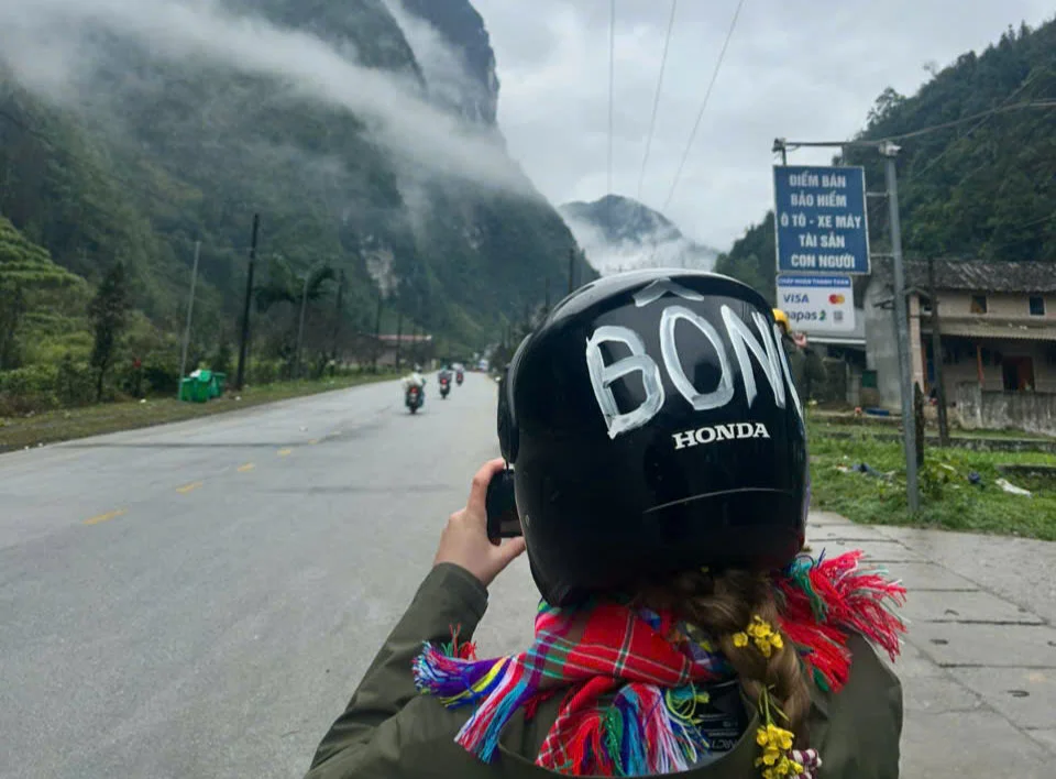 Person in helmet with "BON," photographing misty mountains. Colorful scarf. Motorbikes on a road. Sign in Vietnamese nearby. Overcast mood.