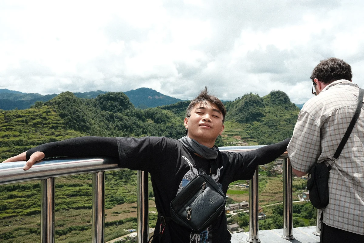 A person poses with arms outstretched on a balcony overlooking green hills, wearing a black outfit and bag; another leans on the railing.